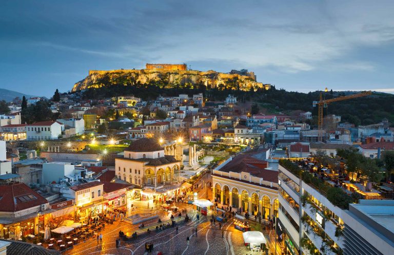 Illuminated Acropolis hill glowing above rooftop restaurants and city lights at dusk