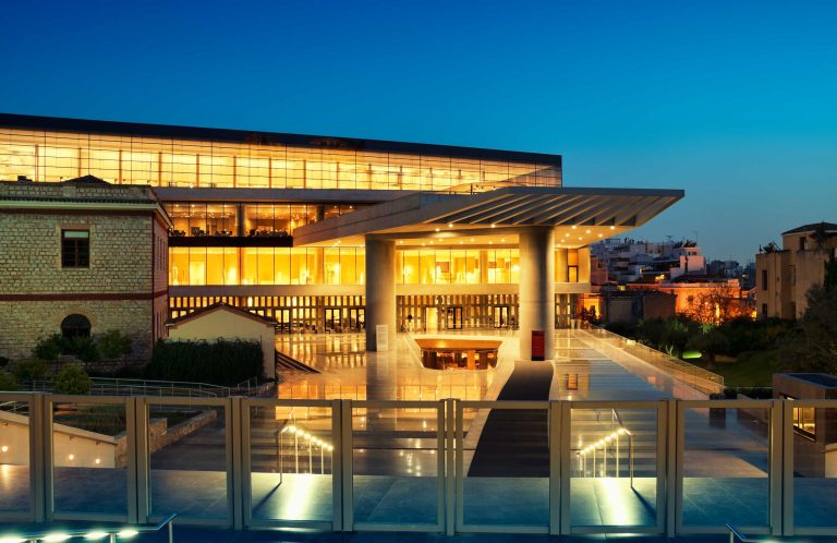 Modern building with glass walls lit from inside at dusk, with glass railings and paving in front
