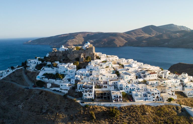 Whitewashed cubic houses climbing a hillside up to a hilltop castle, dark blue sea beyond