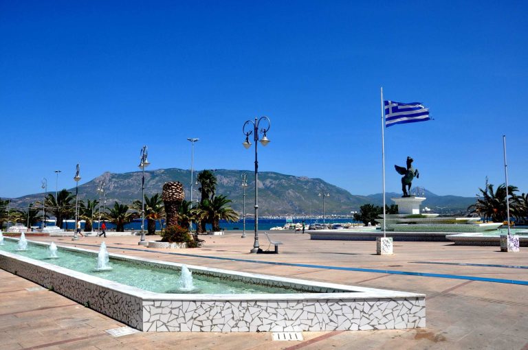 Fountain plaza with a statue of Pegasus, Greek flag, and palm trees, hills in the background