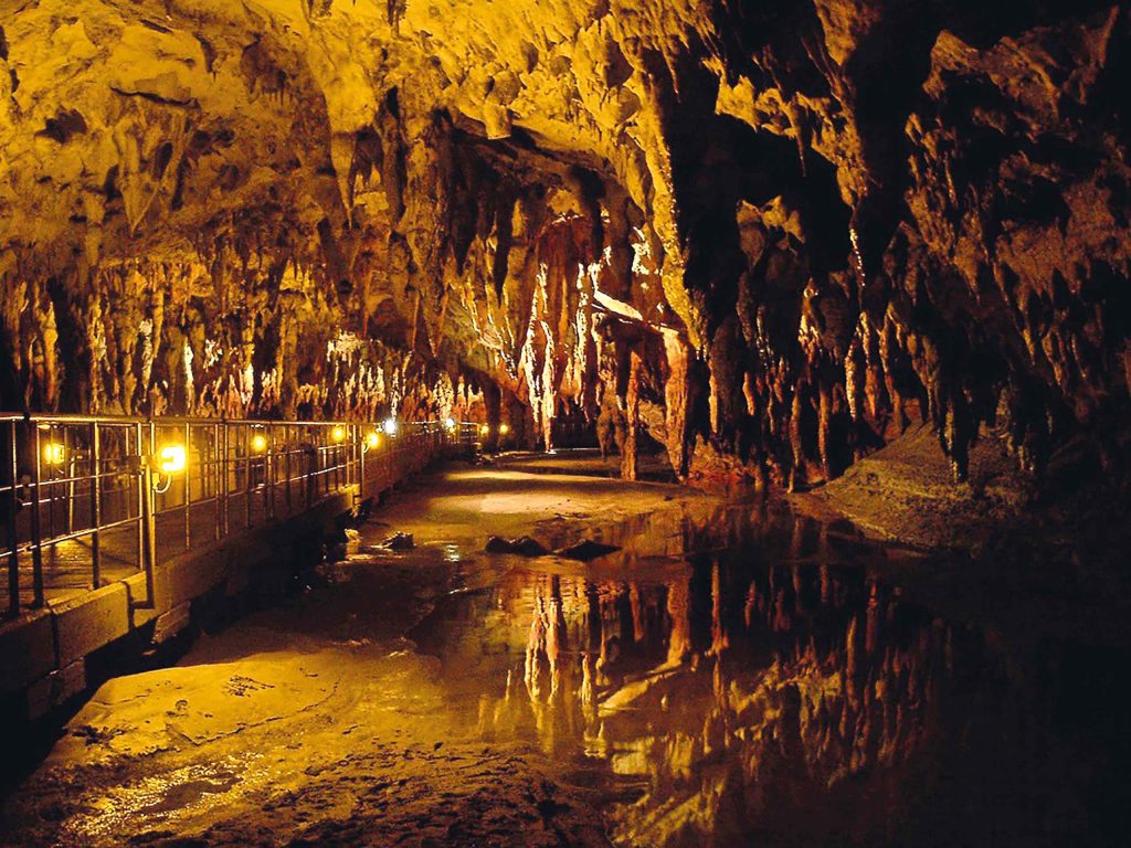 Illuminated interior of Aggitis river springs cave, with a wooden walkway over reflecting water.