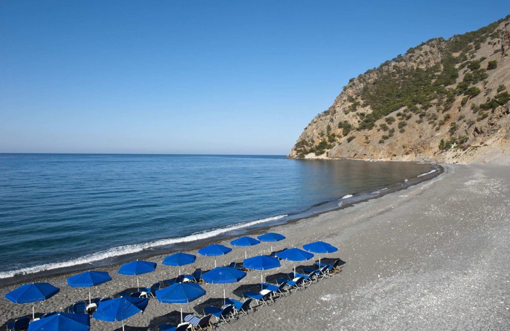 Dark pebble beach with rows of blue sunbeds, and a steep hill at its end
