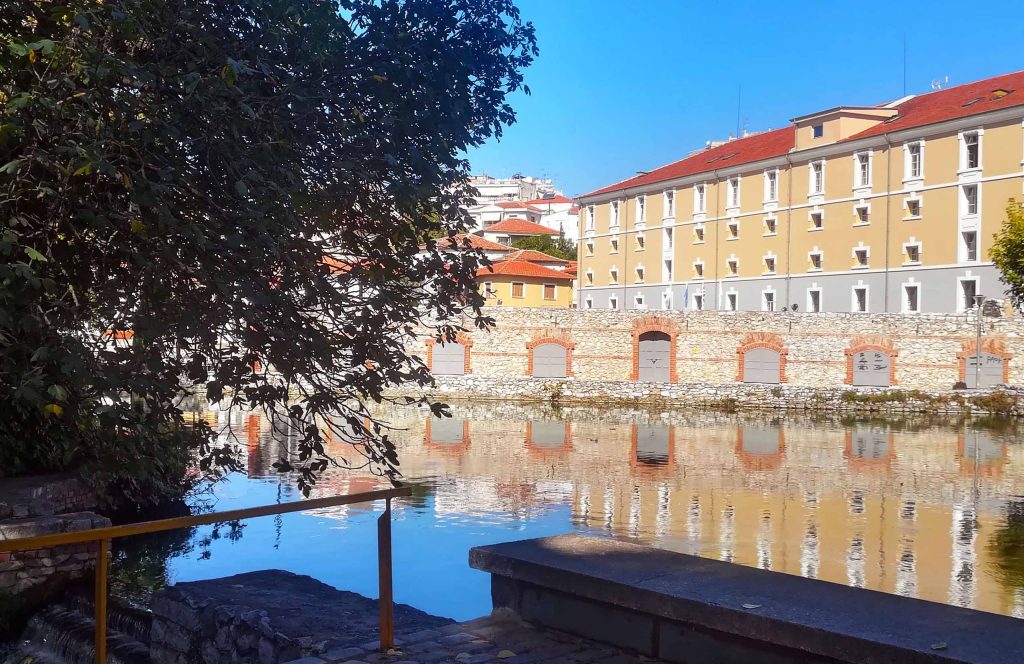 Restored brick tobacco warehouse reflected in still water, with trees along the bank, in Drama.