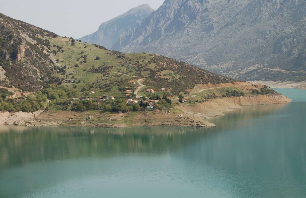 Village houses on a green slope above a lake with low water, with remains of older houses on the bare shore and a rocky mountain behind