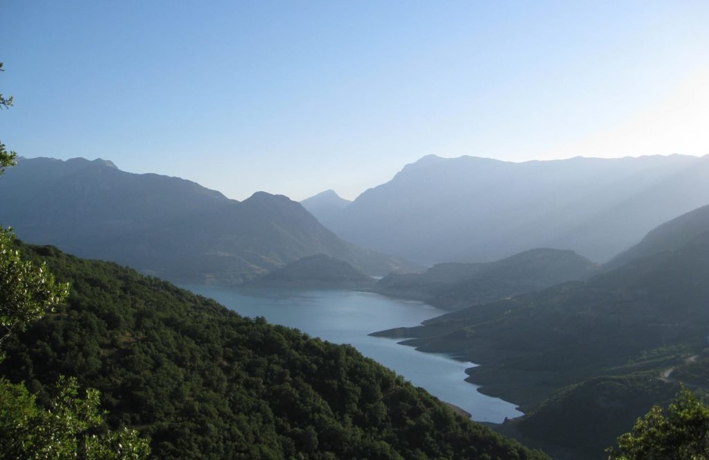 Long narrow arm of a lake between forested slopes, with high mountains in haze behind