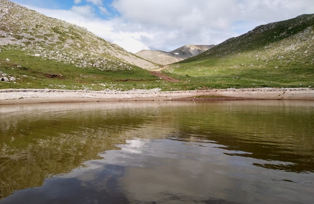 A small lake between grassy slopes on Mount Tymfristos, with clouds reflected in the water