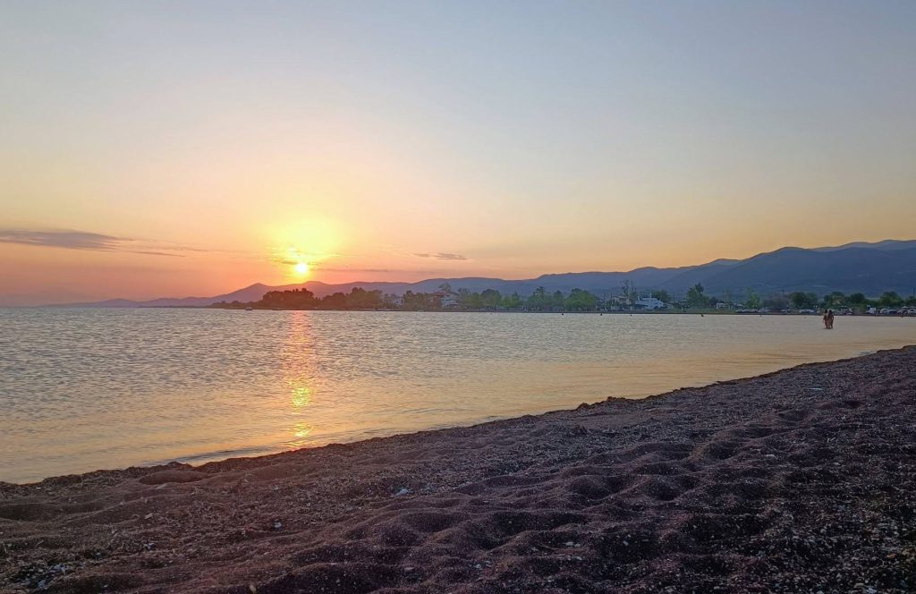 Sun setting over the Maliakos Gulf, with a curve of sand and pebble beach in the foreground