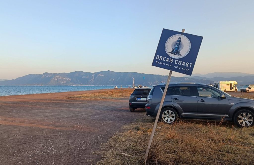 Cars parked on the sand spit of Faros beside a beach bar and kite surf sign, with a kite in the air across the water