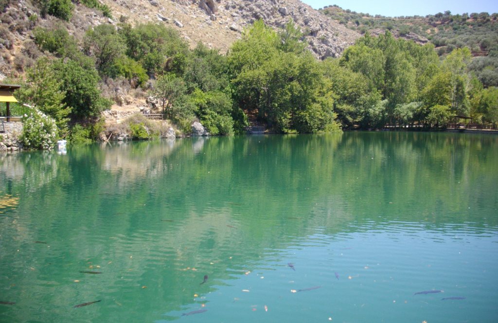 Small lake with green water and trees along its shore below rocky slopes at Zaros