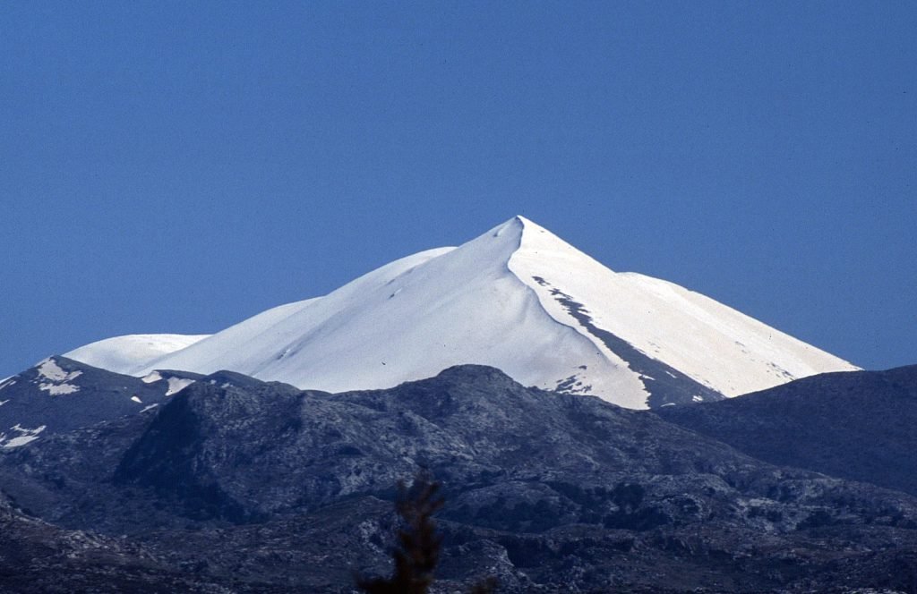 Snow-covered peak of Mount Ida against a deep blue sky, above dark rocky ridges