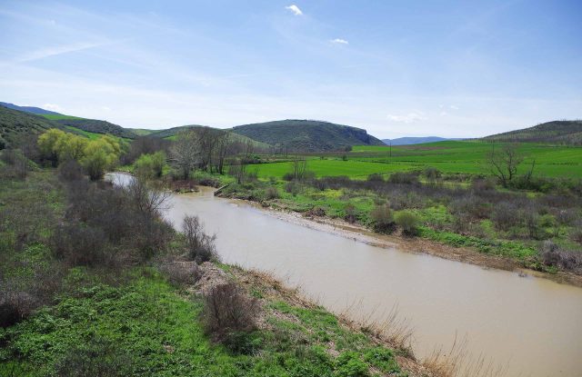 Brown river curving between green banks and low hills, under a blue sky