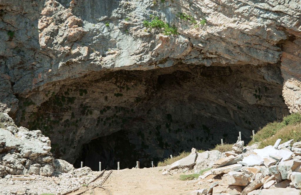 Wide dark mouth of the Idaion Cave under a limestone cliff, with a path leading in