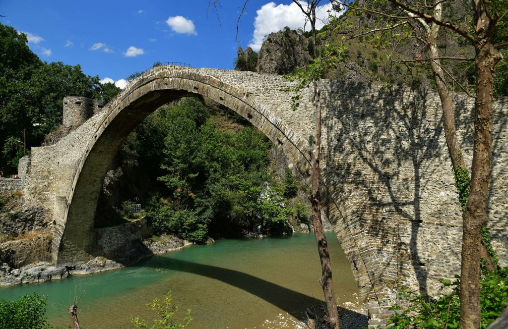 High single-arch stone bridge over green water, with trees and a rocky hill behind