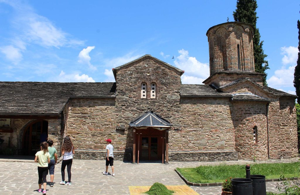 Stone Byzantine church with a tall tower, and visitors walking in the paved courtyard