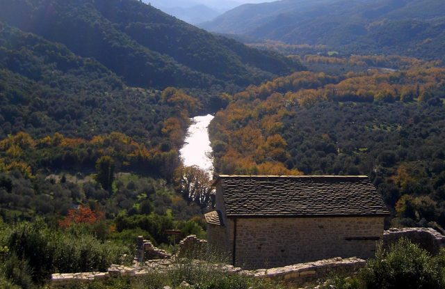 Stone church with a slate roof on a ridge, above a valley where a river winds between trees in autumn colours