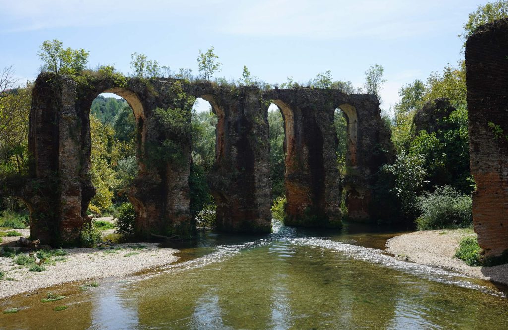 Row of tall brick arches crossing a shallow river with gravel banks, among trees