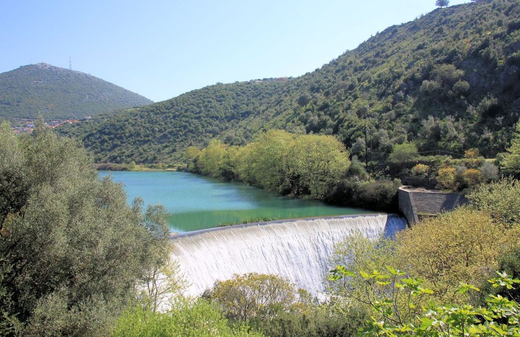Water falling over a curved dam wall below a narrow green reservoir, between wooded hills