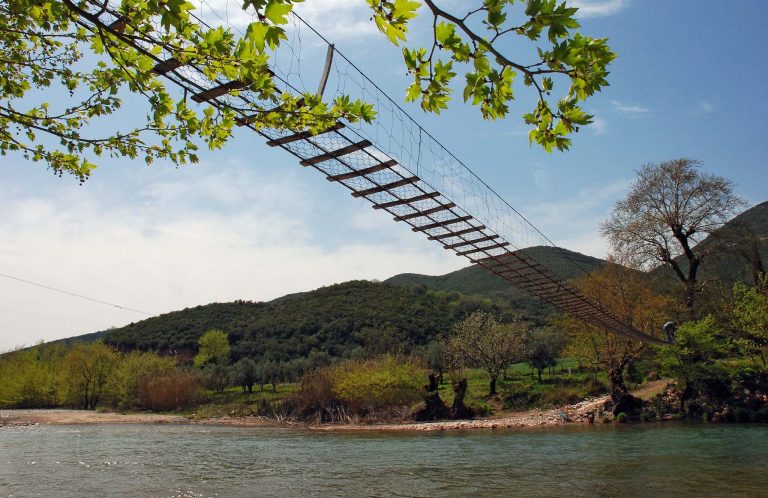 Suspension footbridge crossing high above a river, with green hills behind and tree branches in the foreground