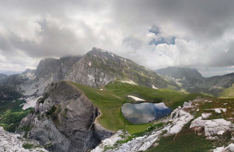 The lake Drakolimni on a green ledge of Mount Tymfi, with the peak of Ploskos above it and cloud over Gamila