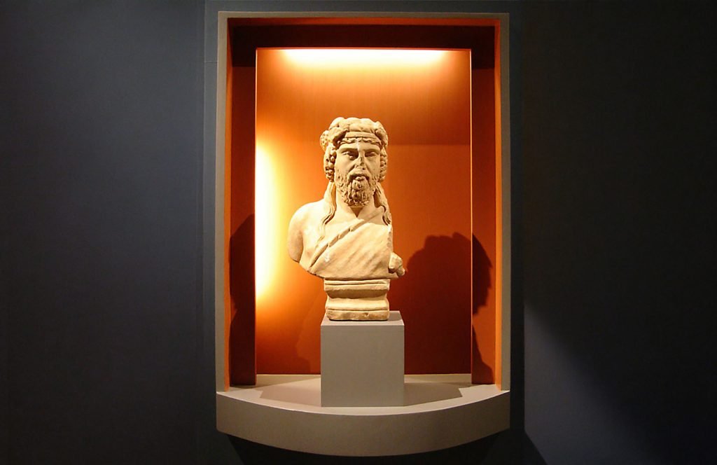 Marble bust of the head of the god Dionysus, displayed in a lit museum case.