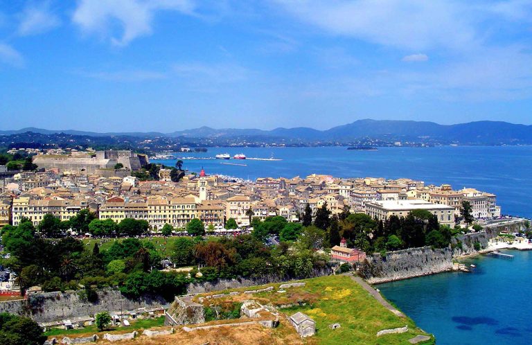 Aerial view of a coastal old town with red-tiled roofs and a fortress, mountains across the water