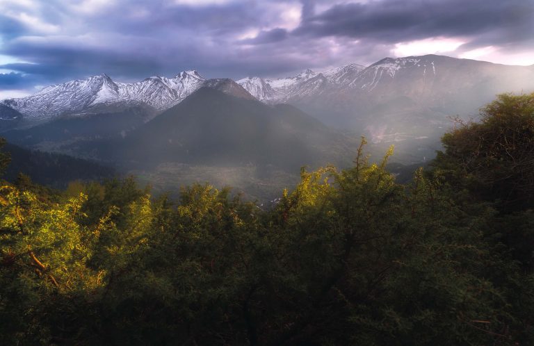Snow-covered peaks of Lakmos under dark clouds, seen over a line of trees lit by the sun