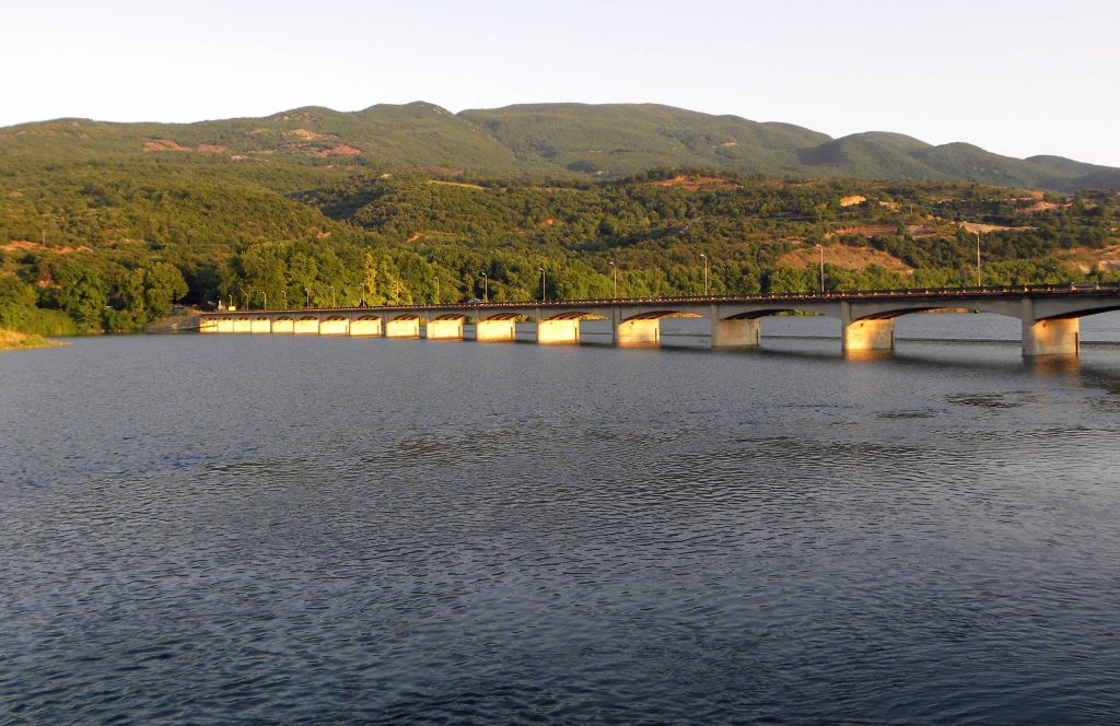 Long road bridge on many piers crossing a calm lake below low wooded hills, in warm evening light