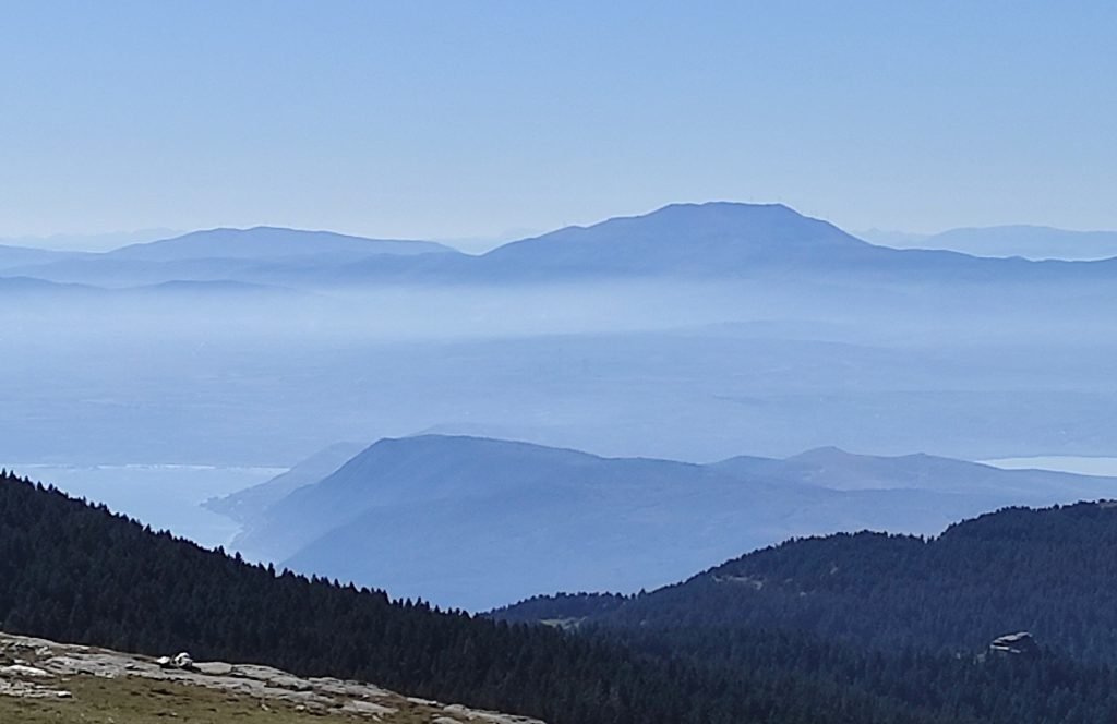 Hazy blue view over Lake Vegoritida and a mountain behind it, seen from above dark forested ridges