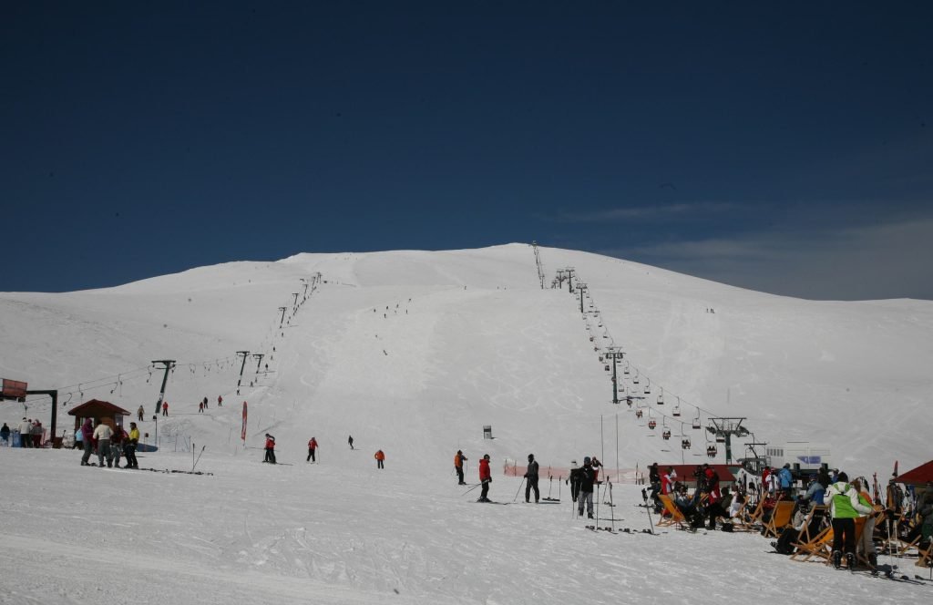 Wide snow-covered slope with two lift lines climbing to a rounded summit, with skiers and people on deckchairs at its foot