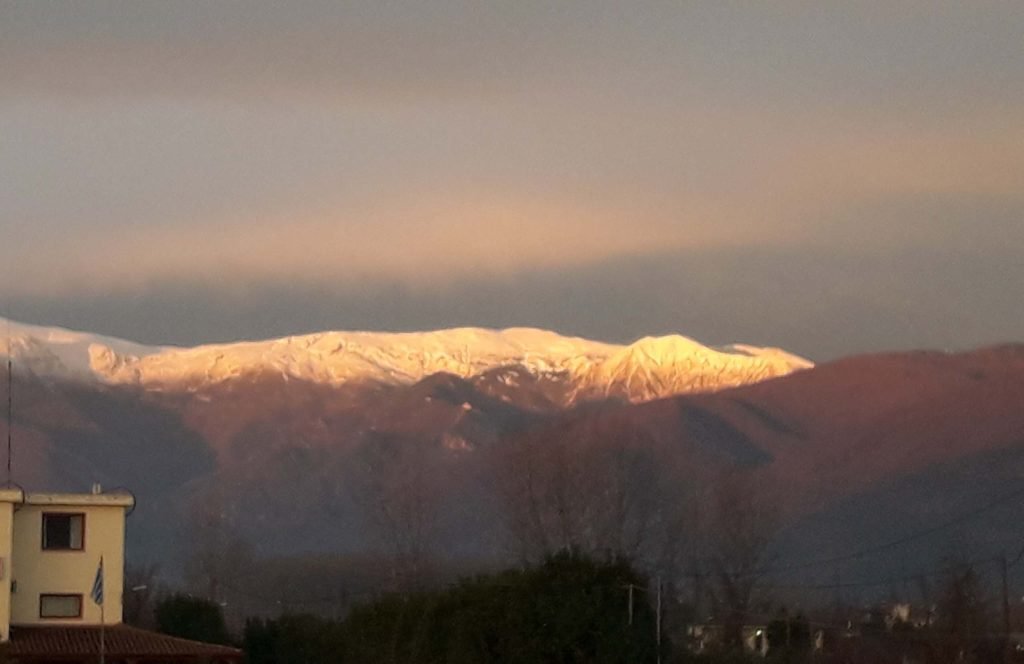 Snow-covered ridge of Mount Voras lit by low sun under a dark cloudy sky, above dark lower slopes, with a building at the lower left