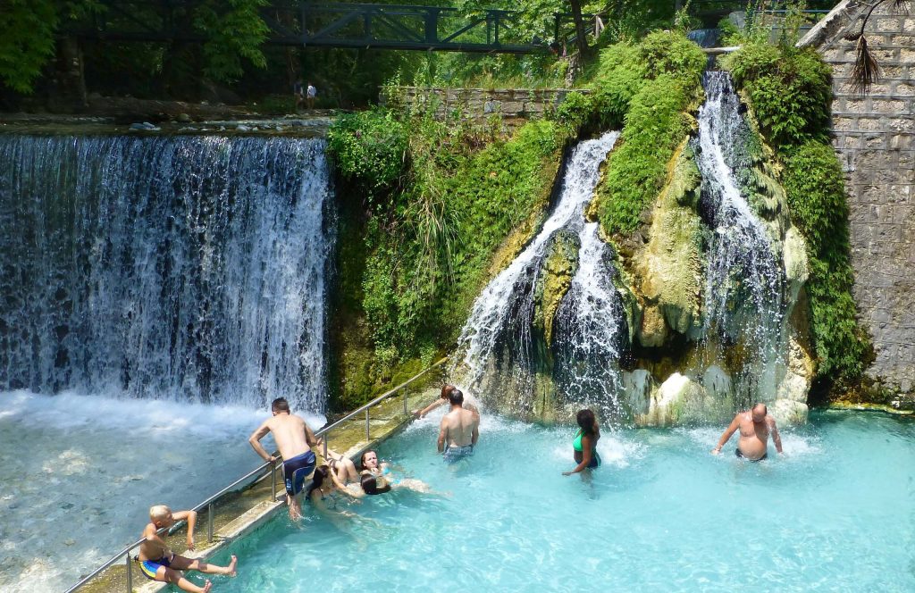 Bathers in a pool of pale blue water below two waterfalls and a rock covered in green plants