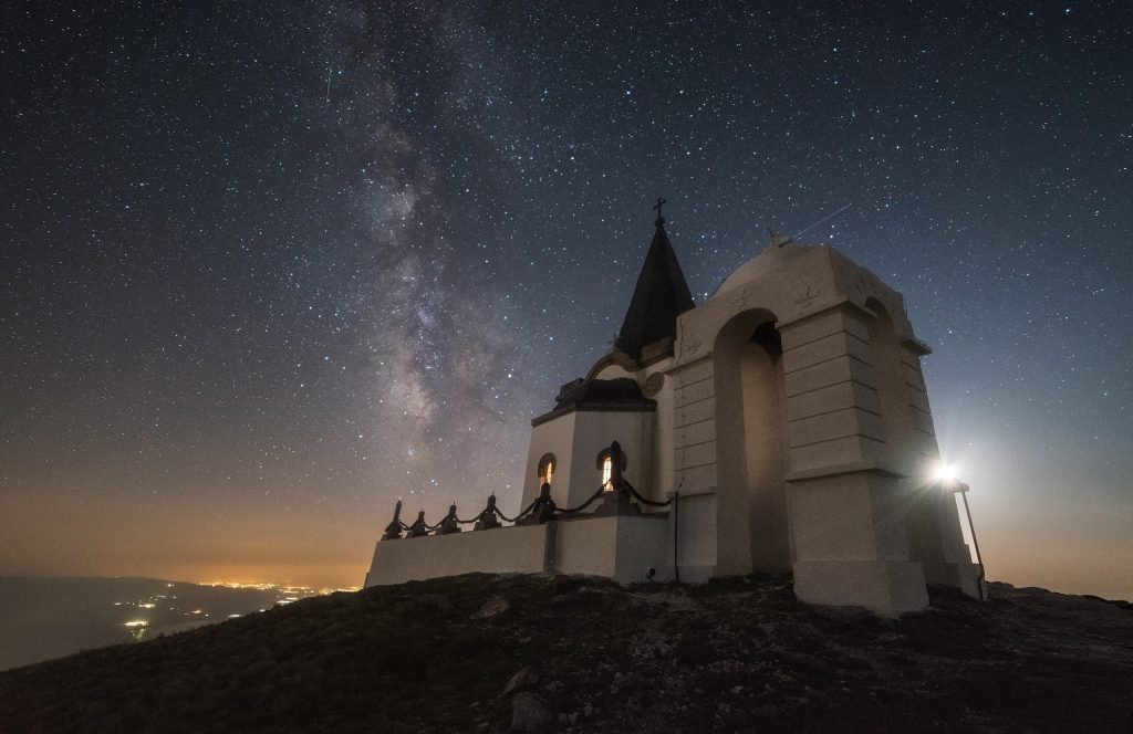 Small chapel with lit windows on a dark hilltop under a sky full of stars and the Milky Way, with town lights far below