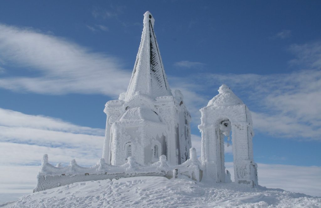 Small chapel with a pointed roof and a bell tower beside it, both completely covered in white ice, on snowy ground
