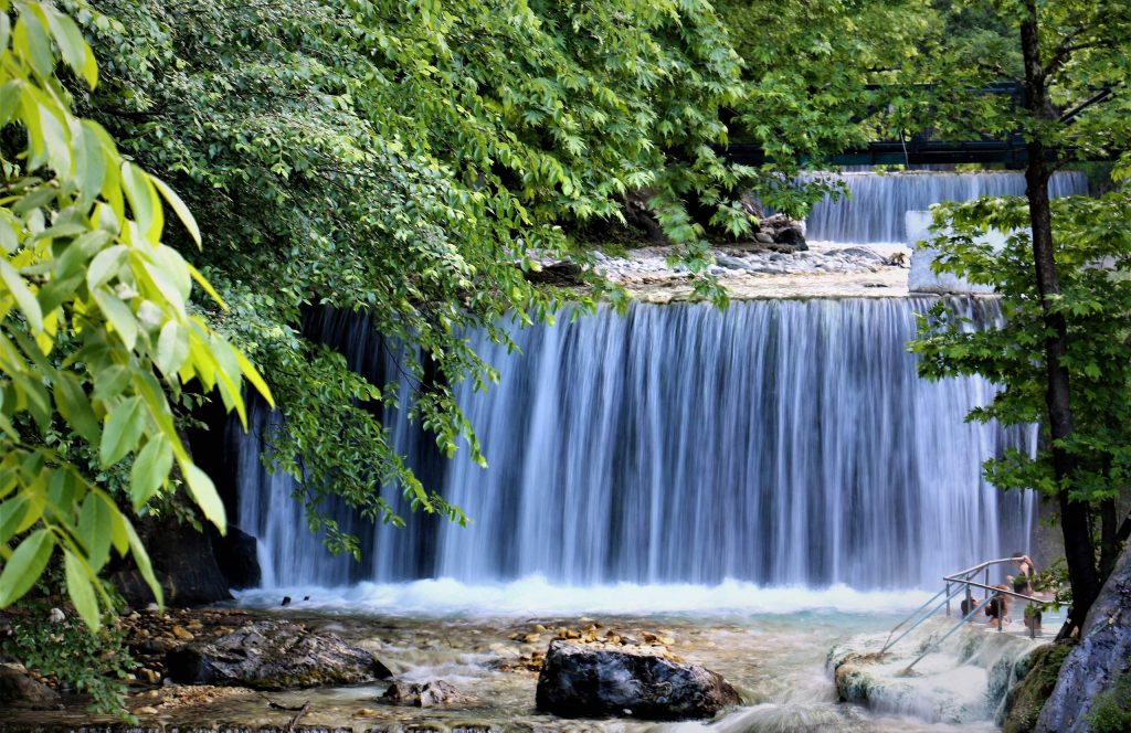 Wide waterfall dropping over a ledge into a rocky stream, framed by green trees