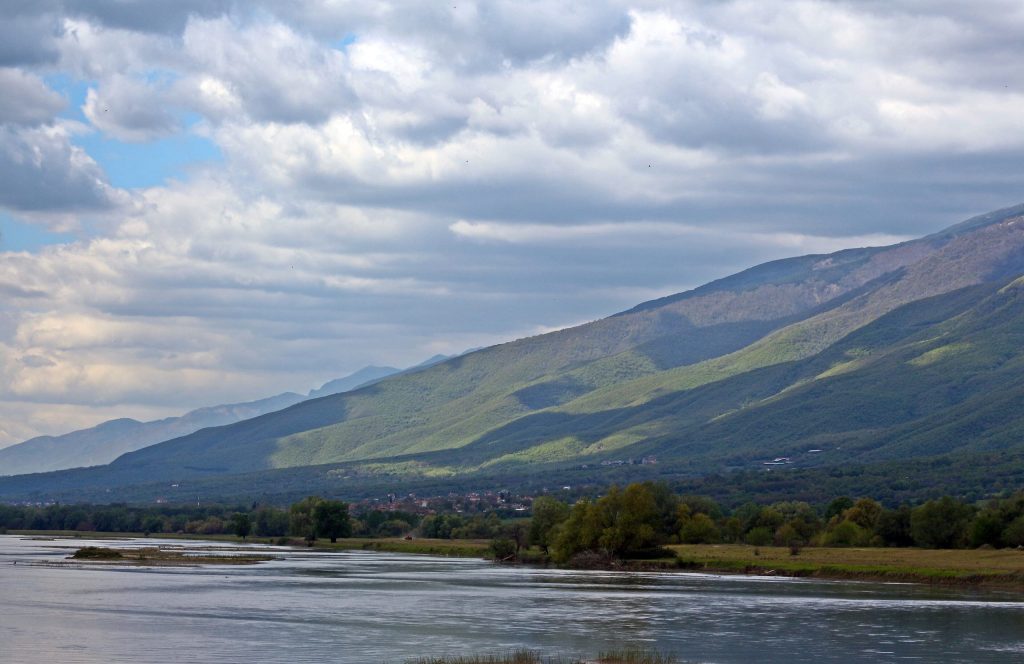 Wide river in the foreground, with trees on the far bank and long green mountain slopes under clouds
