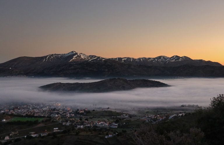 Fog lying over the Lasithi Plateau, with a village below and the snow-streaked peaks of Dikti against an orange sky