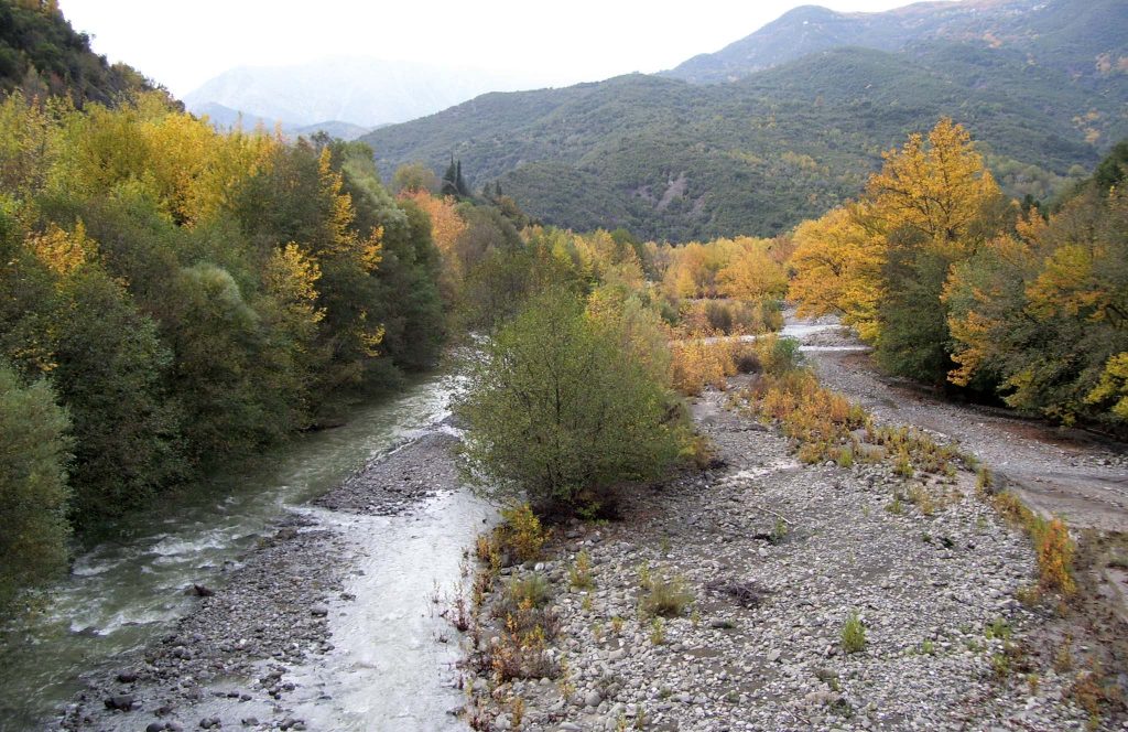 Wide gravel riverbed with a shallow stream, between trees in autumn colours and green mountains