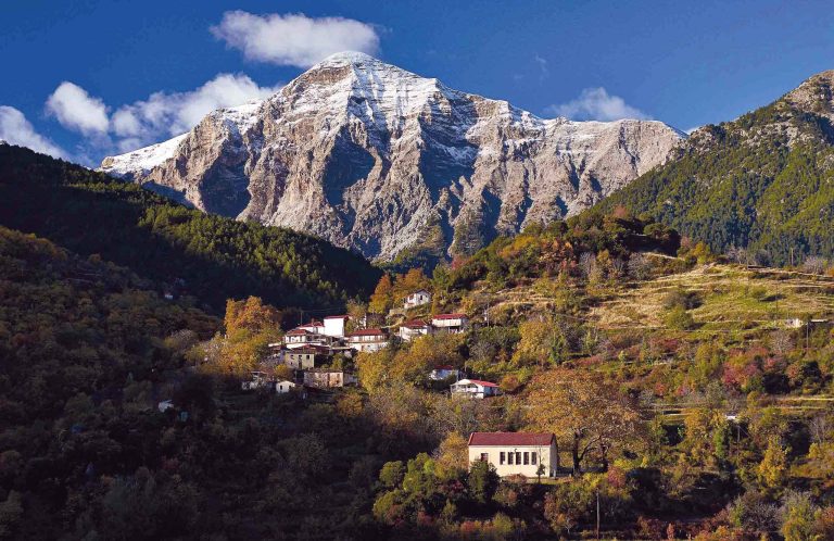 The rocky peak of Neraidorachi with a dusting of snow, rising above a few houses of Mesorougi on a wooded hillside