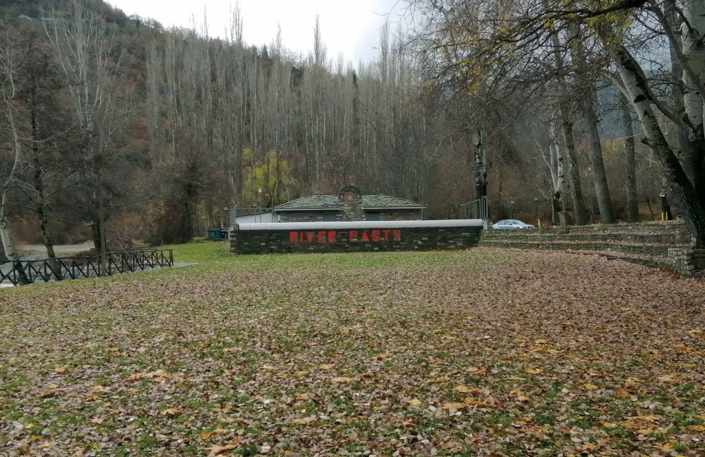 Grassy riverside ground covered with fallen leaves, with a low building and tall bare trees behind