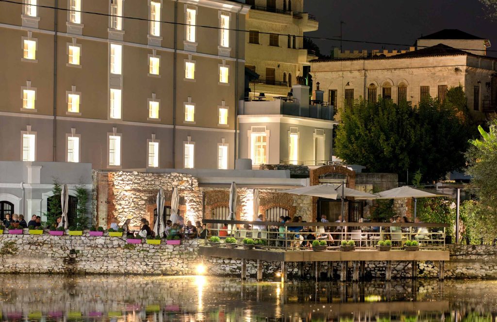 Outdoor dining tables beside a canal, with the illuminated Tobacco Warehouse building in the background at dusk.