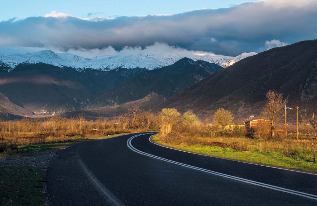 Curving road through a plain with bare orange-brown trees, and the snow-covered peaks of Mount Voras under cloud in the distance