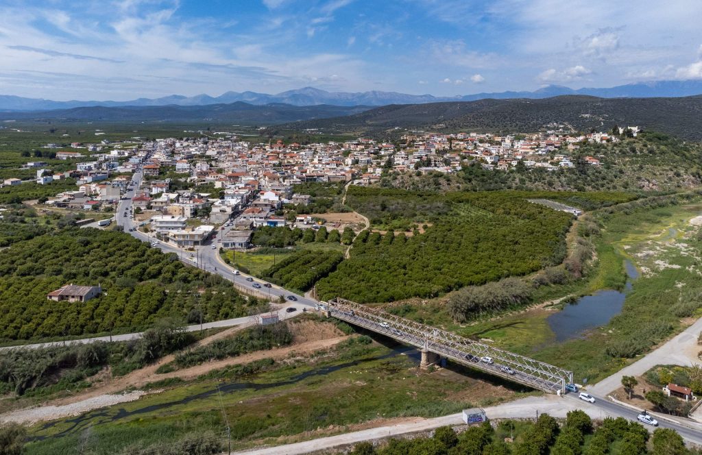 Aerial view of the river passing under a metal road bridge, with the town of Skala among citrus groves behind and mountains on the horizon