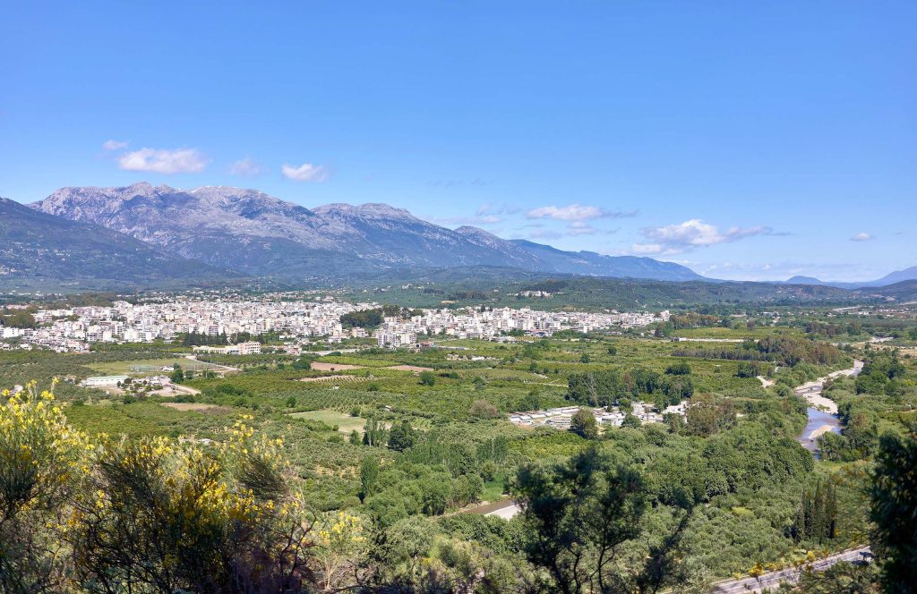 A town spread across a green valley, a river winding on the right, and a long mountain range behind, with yellow shrubs in front