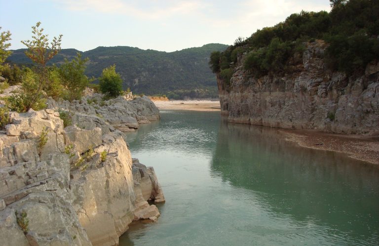 Calm green river between low pale rock walls, with trees on the right bank and hills behind