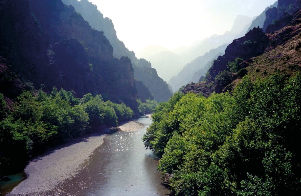 River on a wide gravel bed between steep wooded slopes, with hazy peaks behind