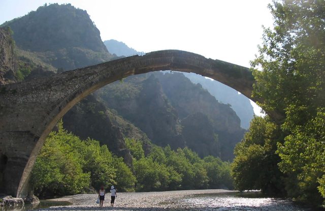 High stone bridge with one arch over a river bed, with two people walking below and mountains behind