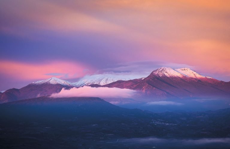 Snow-covered mountain with a rounded main summit under a pink and purple sky, above dark lowland with strips of fog