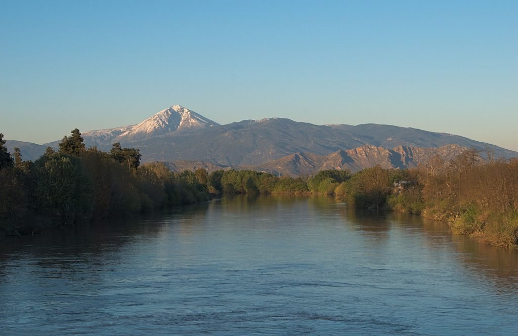 The snow-capped cone of Mount Ossa rising behind the tree-lined Pineios River