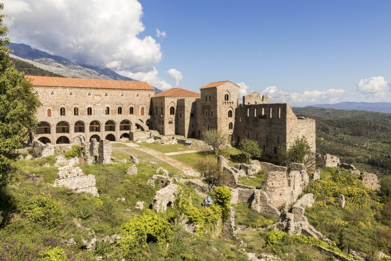 Large stone palace buildings with arched windows on a hillside, with stone walls and bushes in front