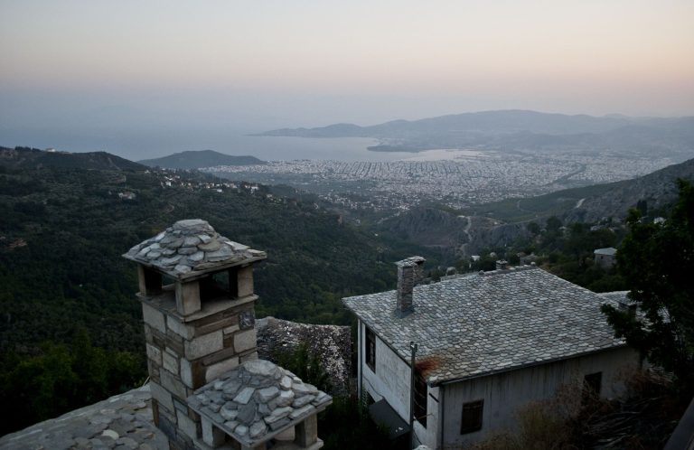 Slate roofs and a stone chimney in the foreground, with the city of Volos and the Pagasetic Gulf far below